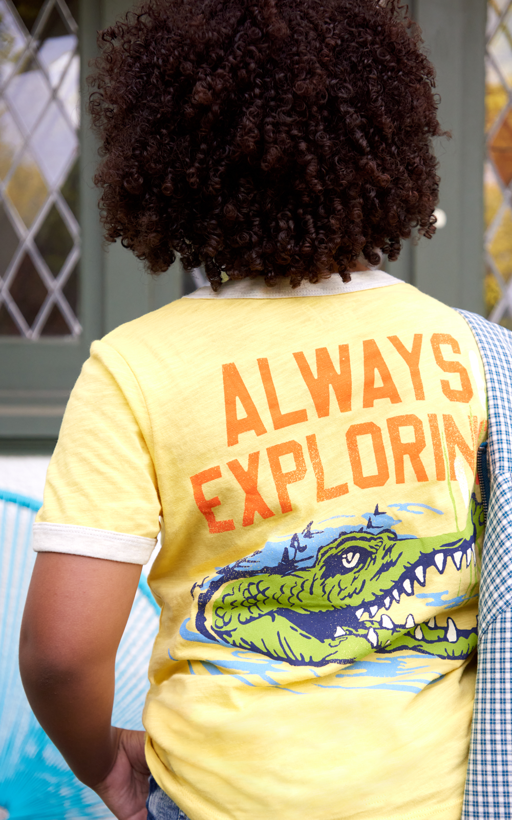 BACK VIEW OF KID WEARING YELLOW T-SHIRT WITH "ALWAYS EXPLORING"