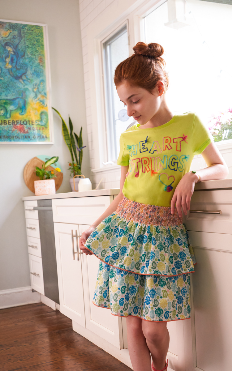 Front view of red headed child wearing a blue and green multi-ruffle skirt and green tee