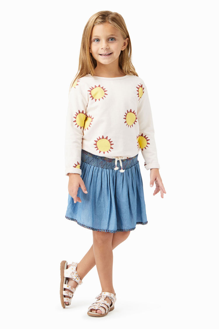 Girl wearing white long sleeve top with yellow sun pattern, blue skirt with colorful thread trim, and daisy print sandals. 