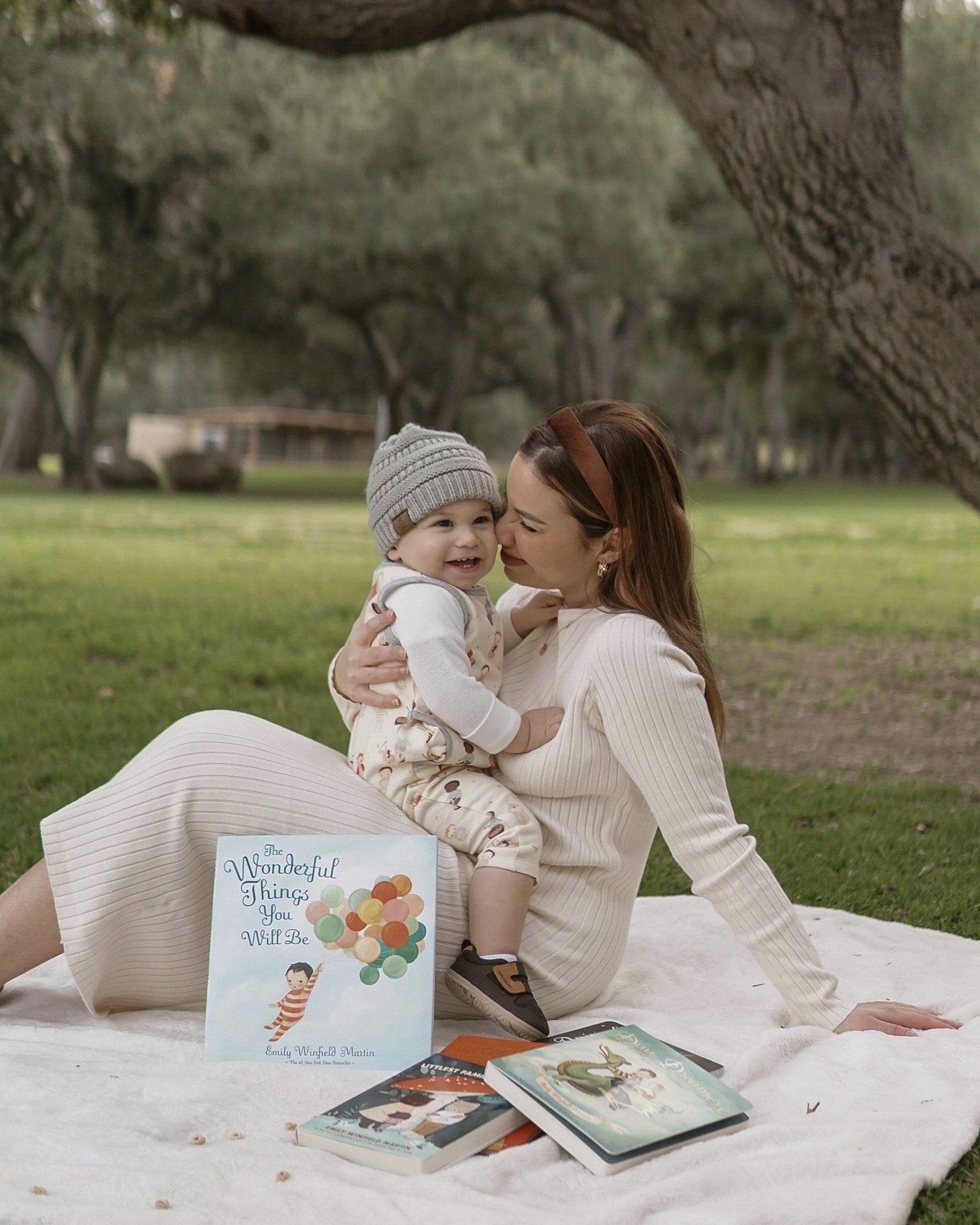 Woman with long dark hair and baby with a gray hat sitting on a blanket with storybooks in a park.